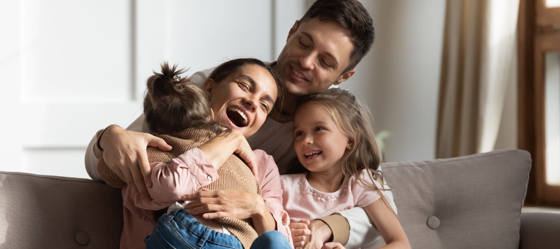 a family of a mom, dad and two young daughters sitting on a couch hugging each other and laughing