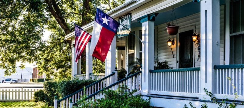 front porch of a house with american and texas flags hanging, surrounded by trees and greenery