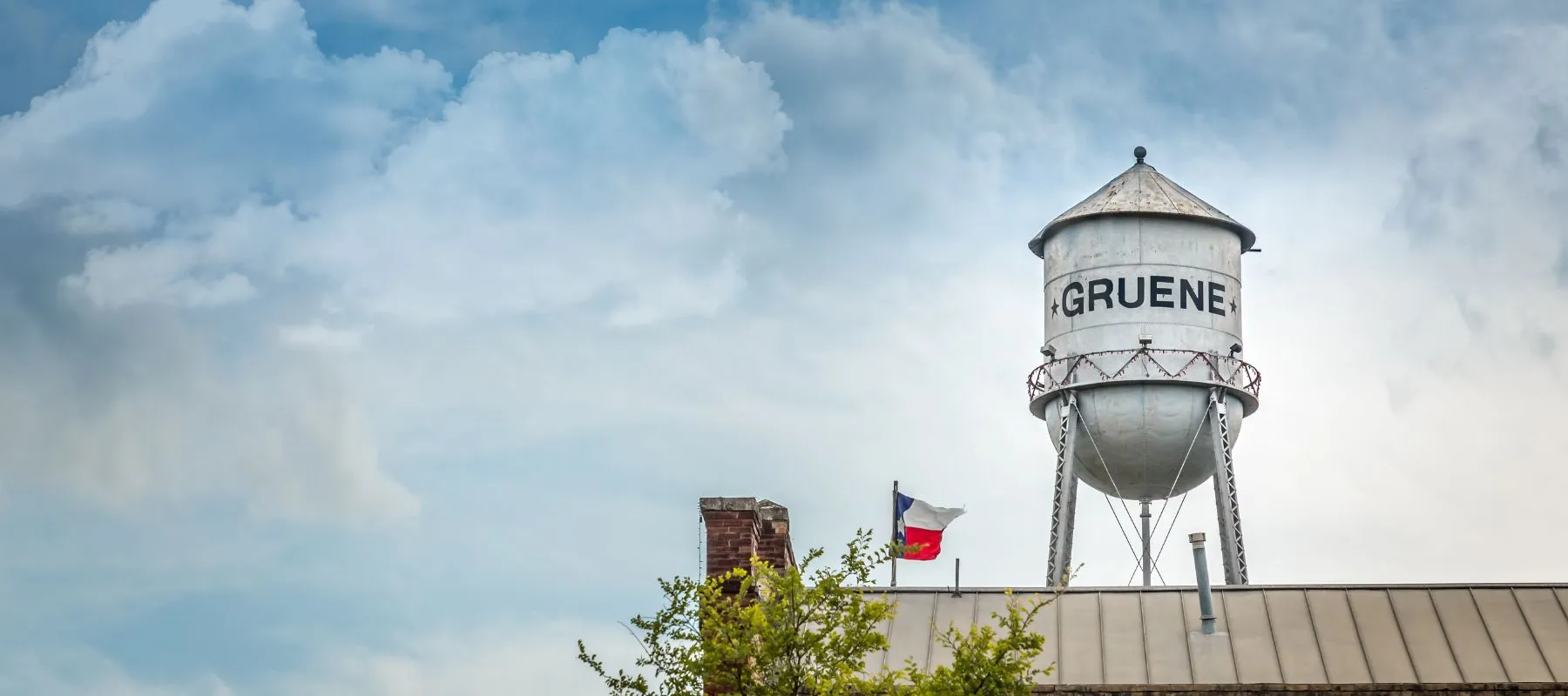 Historic water tower labeled "GRUENE" rises above rooftops and a waving Texas flag under a cloudy sky