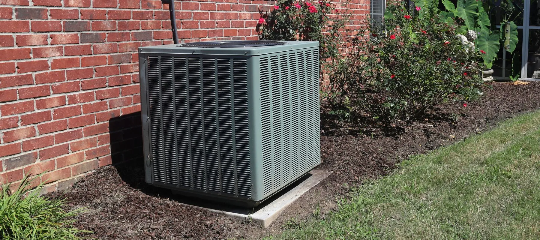 Outdoor air conditioning unit installed next to a red brick house surrounded by flower beds and a grassy yard